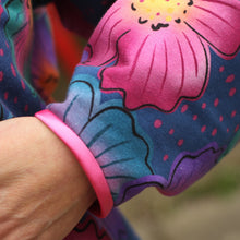 Load image into Gallery viewer, A close-up of a person's hand resting on their lap. The person is wearing a colorful long-sleeve top with a vibrant floral pattern in pink, teal, purple, and blue.