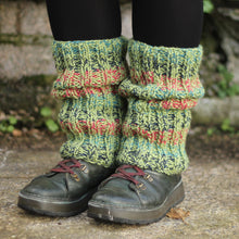 Load image into Gallery viewer, A person wearing chunky knit leg warmers in pesto green, raspberry, ocean and navy blue over black leggings, paired with dark outdoor shoes, standing on a concrete surface with a stone wall in the background.