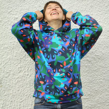 Load image into Gallery viewer, A person wearing a colorful abstract patterned hoodie with blue, green, and orange shapes on a dark background, standing against a textured light gray wall.