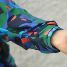 Load image into Gallery viewer, A close-up of a person's hand and wrist wearing a long-sleeve shirt with a bold abstract pattern in green, blue, navy, and red, set against a neutral gray textured background.
