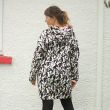 Load image into Gallery viewer, A woman wearing a black and white abstract patterned hooded coat, and black tights, and black boots, standing outdoors in front of a white textured wall