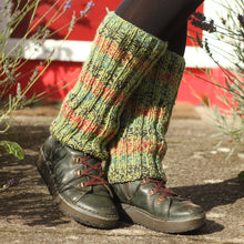 Load image into Gallery viewer, A person wearing chunky knit leg warmers in a pesto green, raspberry, ocean, and navy pattern, paired with black tights and dark lace-up boots, standing outdoors on a stone path with lavender and a red door in the background.
