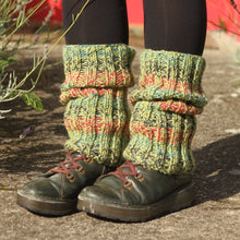 Load image into Gallery viewer, A person standing outdoors on a stone path wearing chunky knit leg warmers in pesto green, with raspberry, ocean, and navy blue stripes, paired with black tights and dark lace-up boots.
