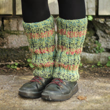 Load image into Gallery viewer, A person wearing chunky hand-knit leg warmers in a variegated pesto green, raspberry, ocean and navy stripe pattern, paired with black tights and dark shoes, standing outdoors against a rustic wooden fence and stone wall.
