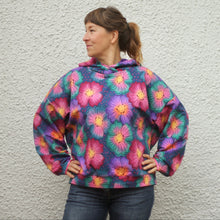 Load image into Gallery viewer, A woman wearing a colorful oversized pullover hoodie with a vibrant pink, purple, and blue floral pattern, standing against a textured white wall.
