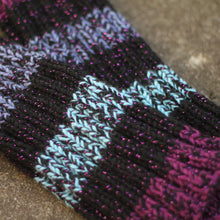 Load image into Gallery viewer, Close-up of a pair of hand-knit wool leg warmers with a striped pattern of black, purple, light blue, and magenta, accented with sparkly purple threads, resting on a textured stone surface.
