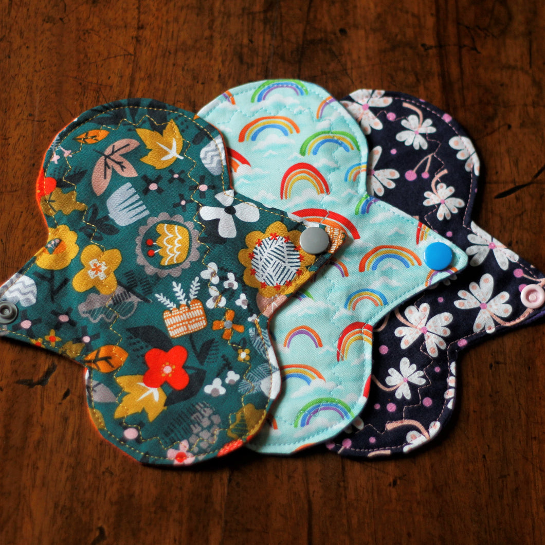 Three pads are laid flat on a wooden table, showing the hibernate, rainbows, and floral on navy patterns.